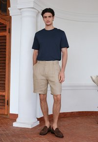 Homme portant une chemise à manches courtes bleu marine, un short beige et des mocassins marron, debout à l'intérieur près d'une colonne blanche et d'une porte en bois.