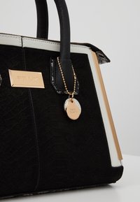 Sac à main noir et blanc avec un motif texturé, fermeture éclair dorée, détails dorés et une étiquette ronde dorée suspendue à une poignée.