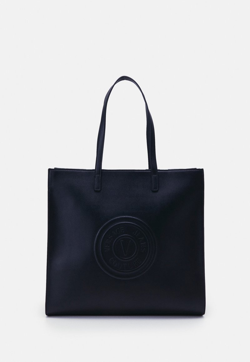 Sac cabas noir avec deux longues poignées et un logo circulaire embossé indiquant Versace Jeans Couture au centre devant.