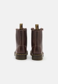 Botas de tobillo de cuero marrón con un suave forro de piel, costuras amarillas y una etiqueta amarilla en la parte trasera. Suela de goma negra gruesa y texturizada.