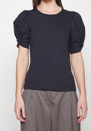 Kvinde iført en mørk marineblå kort top med puffærmer kombineret med løstsiddende taupe bukser, vist fra nakke til hofter på hvid baggrund.