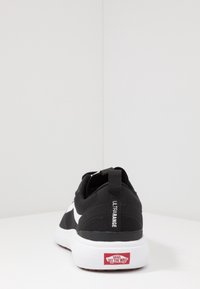 Black athletic shoe with a textured upper, white logo detail, and padded collar. Features a thick white outsole with red accents.