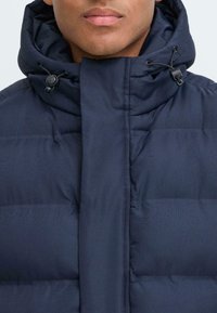 Marineblaue Daunenjacke mit hohem Kragen, verstellbarem Kordelzug und gestepptem Design für Isolation. Vorderer Verschluss mit Knöpfen.