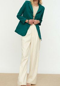 Sininen bleiseri mukautetulla leikkauksella, yhdistettynä kermaan vaaleisiin leveälahkeisiin housuihin, joissa on sileä pinta ja minimalistinen muotoilu.