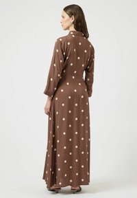 Brązowa sukienka maxi z długimi rękawami, ozdobiona dużymi kremowymi groszkami. Tkanina ma gładką fakturę i lekko opływowy krój.