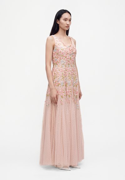 Femme portant une robe rose sans manches, longue jusqu'au sol, avec une broderie florale multicolore et une superposition transparente à pois, debout sur un fond blanc.