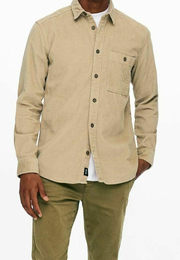 Mann in einem beigefarbenen Kordhemd mit Knopfleiste und Brusttasche über einem weißen T-Shirt, kombiniert mit olivgrünen Hosen.