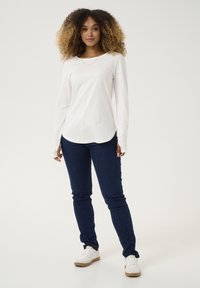 Weißes Langarmhemd mit abgerundetem Saum, kombiniert mit blauen Slim-Fit-Jeans und weißen Sneakers, stehend vor einem schlichten Hintergrund.