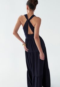 Tumši zila kleita ar mezglotu muguras dizainu, fakturētu audumu, izplešamu apakšmalu un kārtainu svārku. Akcentēta ar gludu jostasvietu un minimālu aparatūru.