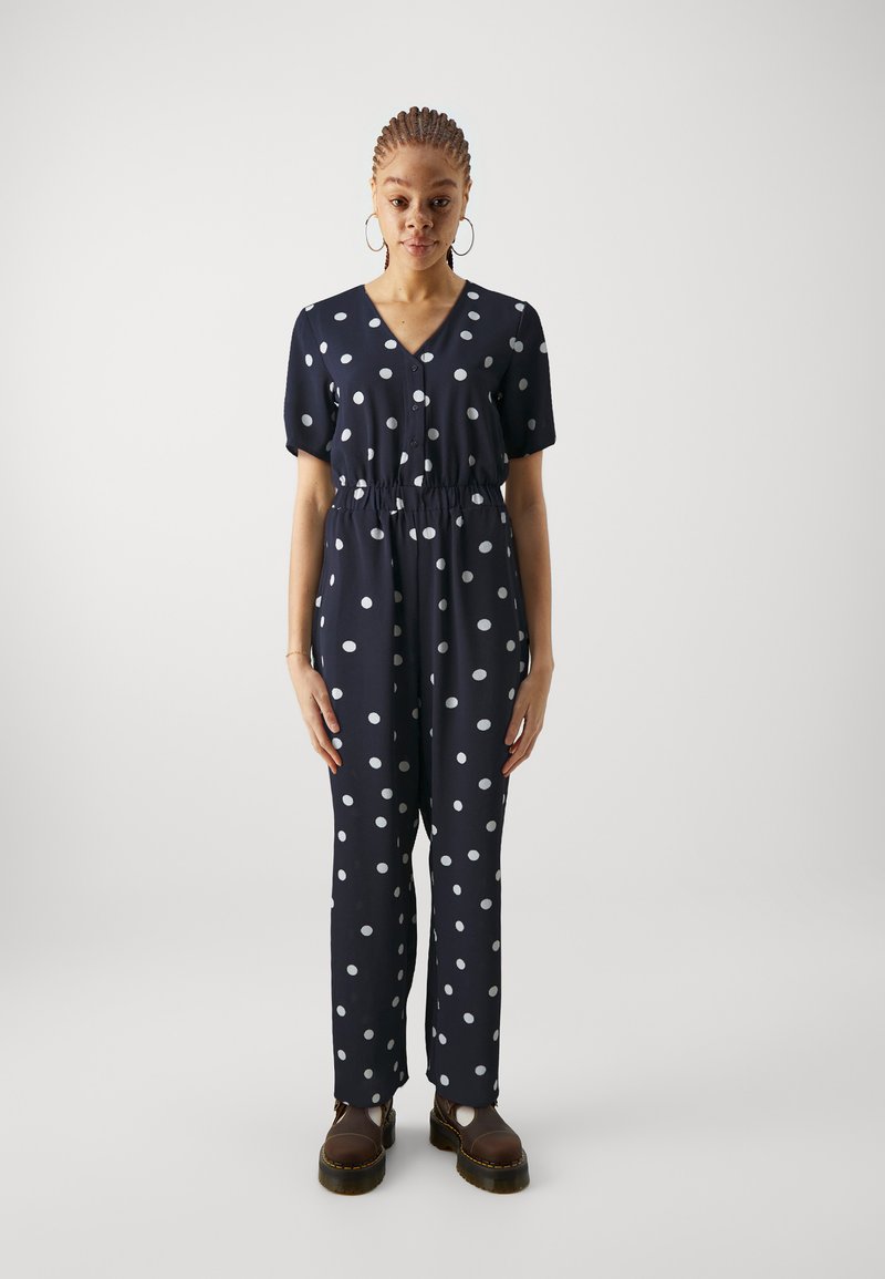 Navy polka dot jumpsuit with short sleeves, V-neck, elastic waist, and wide legs. Features large white dots and front button detail.