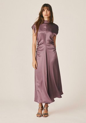 Mujer con un vestido largo sin mangas, de satén malva, con cuello alto, laterales fruncidos y sandalias metálicas de tacón alto con tiras, de pie sobre un fondo liso.