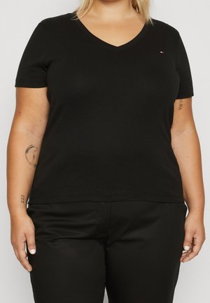 Person wearing a black V-neck short sleeve shirt and black pants, showing tattoos on both forearms.