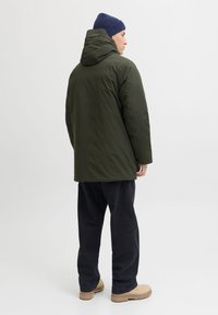 Mees, kes kannab tumerohelist kapuutsiga talvejopet, tumesinist mütsi, musti pükse ja beeže saapaid, seisab küljepealt valge taustaga ees.