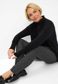 Zwarte cardigan met geribbelde manchetten, gedragen over een zwarte coltrui. Grijs gemusterde broek en zwarte schoenen. Model zit. Minimalistische achtergrond.
