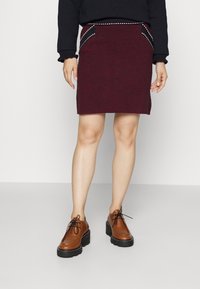 Burgundská pletená sukně s bočními kapsami, zdobená bílým švem. V kombinaci s hnědými šněrovacími botami s tlustou černou podrážkou.