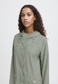 Giacca impermeabile verde chiaro con cappuccio regolabile, bottoni a pressione e tasca frontale, caratterizzata da una texture liscia e lucida.