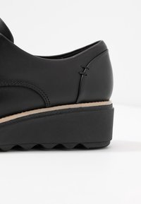 Chaussure en cuir noir avec une semelle compensée basse, une finition texturée et un détail de couture latérale. La semelle extérieure présente un motif ondulé pour l'adhérence.