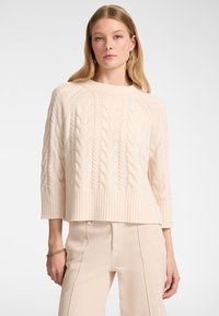 Maglione crema lavorato a maglia con fantasie a treccia, vestibilità comoda e orlo a coste. Abbinato a pantaloni chiari e gioielli minimal.