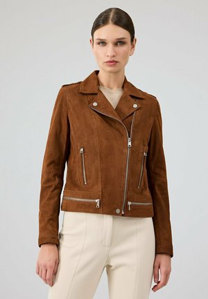 Femme portant une veste de motard en daim marron avec des fermetures éclair argentées et un pantalon de couleur crème, debout devant un fond gris clair uni.