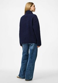 Mörkblå fuskpälsjacka med hög krage, fyrkantig form och långa ärmar, matchat med ljus tvättade vida jeans och beige skor.