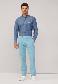 Blauw overhemd met lange mouwen, voorzien van een logo, gecombineerd met lichtblauwe broek en witte sneakers. Glad textuur overal.
