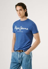 Tricou din bumbac albastru cu mâneci scurte, având inscripția albă "Pepe Jeans London", asortat cu blugi decolorati albaștri și o curea maro.