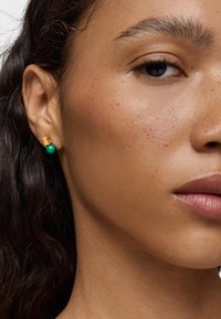Pendientes de stud en tono dorado con forma de oso y una perla verde debajo, sobre una piel con textura natural y luz.