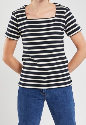 Frau trägt ein kurzärmeliges Oberteil mit schwarzen und weißen horizontalen Streifen, quadratischem Ausschnitt und blauer Jeans vor einfarbigem Hintergrund.