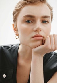 Vista cercana de una persona con pelo corto, vistiendo una parte superior de cuero negro y pendientes de aro dorados, apoyando la barbilla en la mano. Piel suave y fondo claro.