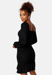 Czarna sukienka z długim rękawem wykonana z teksturowanej tkaniny. Posiada kwadratowy dekolt, marszczone boki oraz falbaniaste wykończenie dołu.