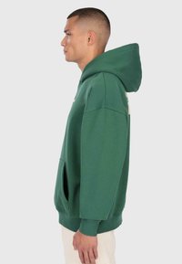 Homme avec les cheveux courts portant un sweat à capuche vert et un pantalon de couleur claire, se tenant de profil contre un fond uni.