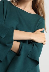 Tyrkysově modrý pletený svetr s širokým výstřihem a objemnými zvonovými rukávy. Měkká textura s žebrovanými manžetami. Univerzální a pohodlný střih.