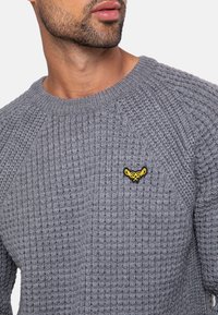 Grijze gebreide trui met een gestructureerd patroon, voorzien van een ronde hals en een klein geel embleem op de borst.