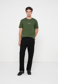 Νέος άνδρας που στέκεται, φορώντας πράσινο μπλουζάκι Tommy Hilfiger, μαύρο παντελόνι και μαύρες μοκασίνες, μπροστά σε απλή λευκή επιφάνεια.