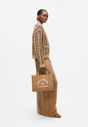 Tan knitted sweater with black pattern, paired with tan wide-leg trousers. Holding a brown tote bag with white text and animal print shoes.