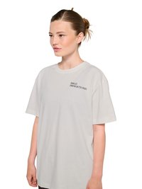 Jeune femme à la peau claire, cheveux attachés, portant un t-shirt gris clair oversize avec le texte noir "HALO 196819721981" sur la poitrine gauche.