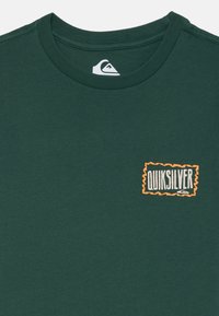 T-shirt verde escuro em algodão, com um logotipo texturizado em laranja e branco, com decote redondo e mangas curtas.