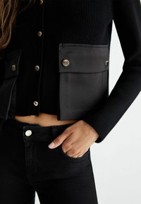 Cardigan nero a costine con due grandi tasche frontali, bottoni dorati e un design corto, abbinato a jeans neri e una mano in tasca.