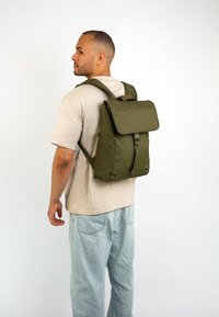 Olívazöld hátizsák hajtható fedéllel, állítható pántokkal és cipzáras zsebbel az elején. Tartós anyagból készült, sima textúrával.