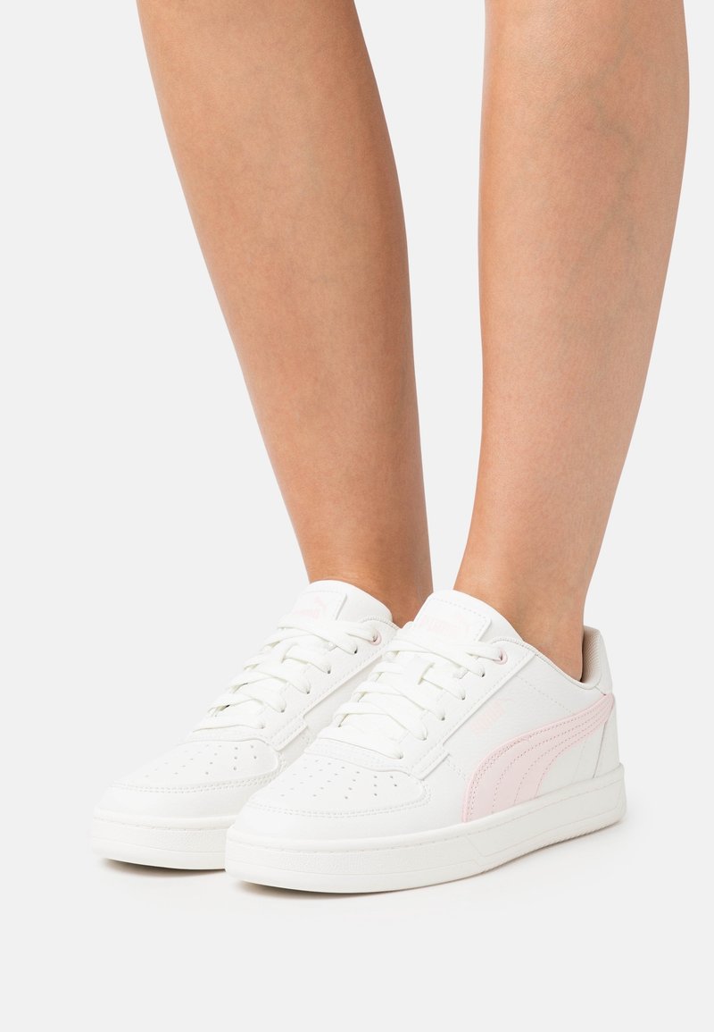 Baskets blanches basses avec des bandes latérales rose clair portées pieds nus, présentées sur un fond blanc uni.