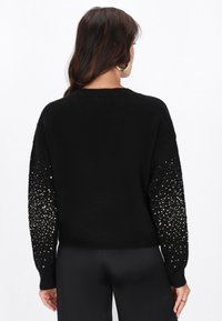 Zwarte sweater met een zachte textuur, een losse pasvorm en paillettenaccenten op de mouwen, wat een opvallend visueel contrast creëert.