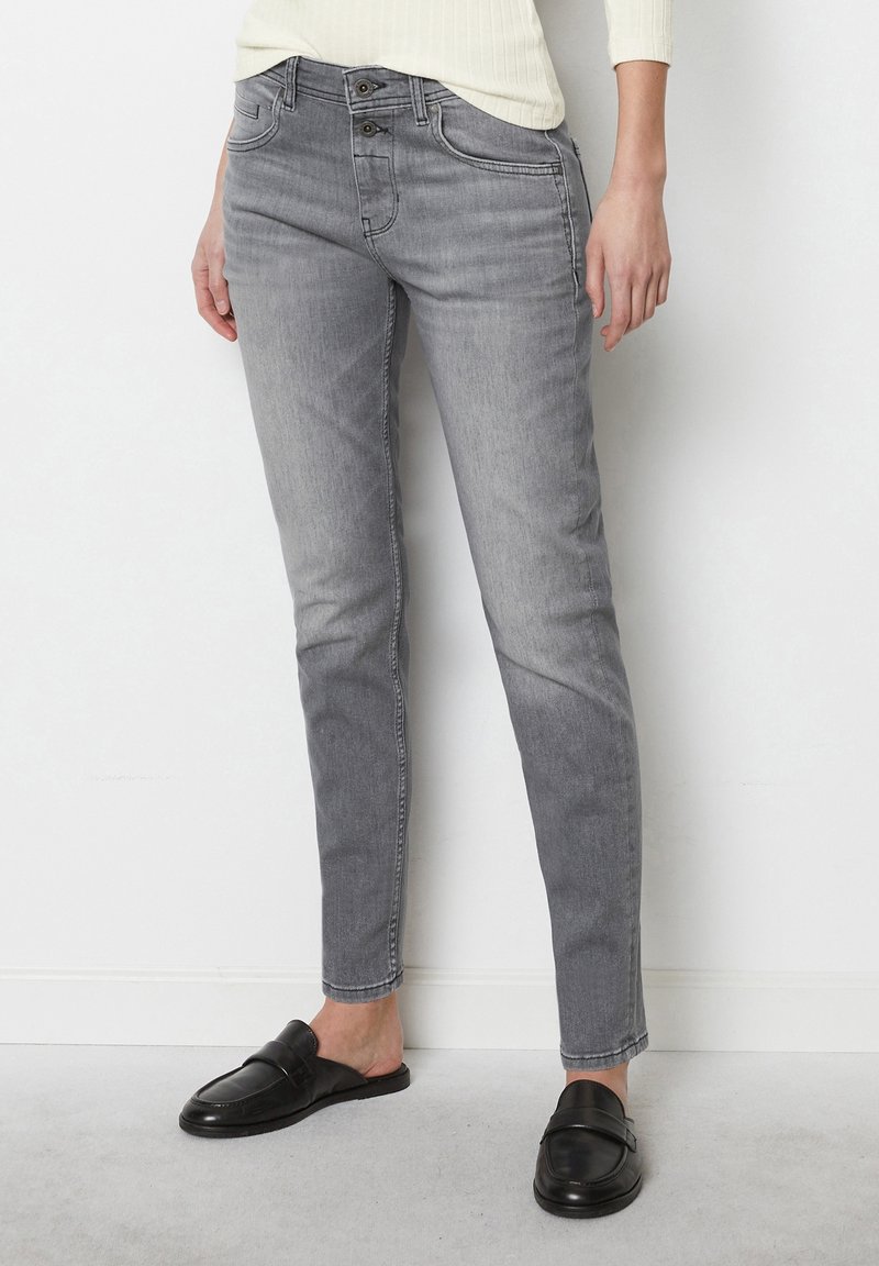 Personne portant un jean gris ajusté avec taille à double bouton, haut blanc cassé, et mocassins noirs sans lacets, debout contre un mur blanc.