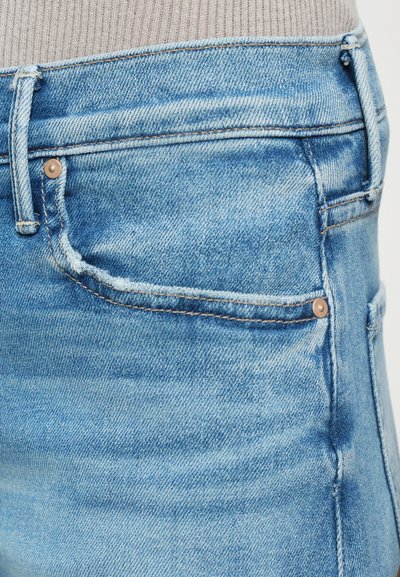 Close-up of faded blue denim jeans with visible stitching, belt loops, and a front pocket, worn with a ribbed beige top.