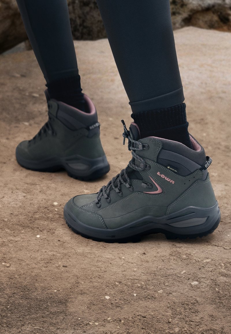 Personne portant des chaussures de randonnée grises et roses avec des chaussettes noires et des leggings foncés, debout sur un sol rocheux.