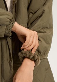 Vihreä vuorattu takki, jossa on joustavat hihansuut; hihansuidemme karvavärityksessä on yksityiskohtia. Käsivarsi on asetettu lähelle takin avointa kohtaa, paljastaen materiaalin rakenteen.