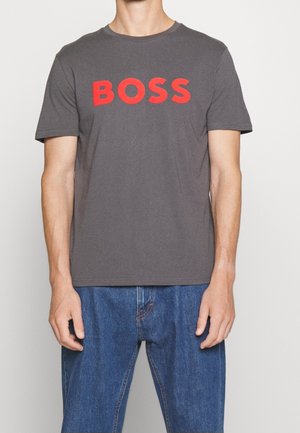 Hombre con una camiseta gris de manga corta con la palabra "BOSS" en grandes letras rojas en el pecho y vaqueros azules.