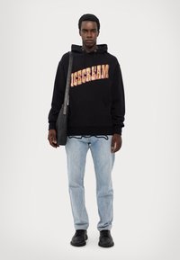 Schwarzer Hoodie mit "EISCREME" in bunten Buchstaben, kombiniert mit hellblauen Jeans und schwarzen Schuhen, dabei eine schwarze Umhängetasche.