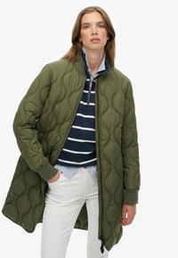 Cappotto trapuntato verde oliva con colletto alto, caratterizzato da una chiusura con zip, polsini a coste e una siluetta leggermente allungata. Indossato sopra una camicia a righe.