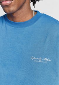 Sinivihreä puuvillapaita, jossa on pyöreä kaula-aukko, ja vasemmassa rintataskussa valkoisella brodeerattu teksti "Rétro-nité Atelier Création de l'Art".