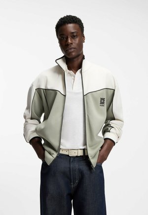 Młody mężczyzna w białej i szarej kurtce na suwak z logo "HUGO", białej koszulce polo, ciemnych dżinsach i białym pasku, z rękami w kieszeniach.